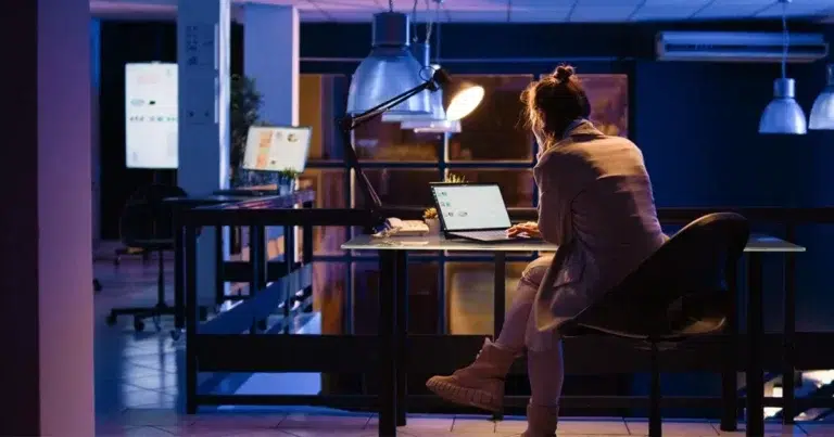 Nachtzuschlag: Eine Frau sitzt im Büro an einem Schreibtisch am Laptop. Nur eine Schreibtischlampe ist an, sonst ist alles dunkel. So kann eine Nachtschicht inkl. Nachtzuschlag aussehen.