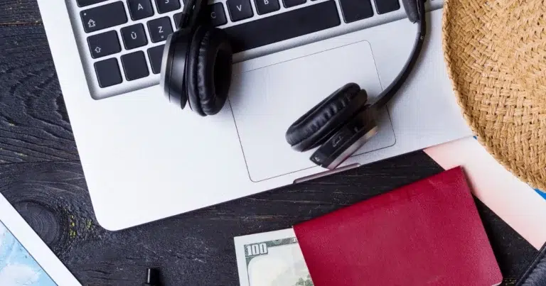 Ein Laptop mit Kopfhörern liegt auf einem Tisch neben einem Reisepass, Bargeld und einem Strohhut und symbolisiert die Vorbereitung auf eine Urlaubsreise.