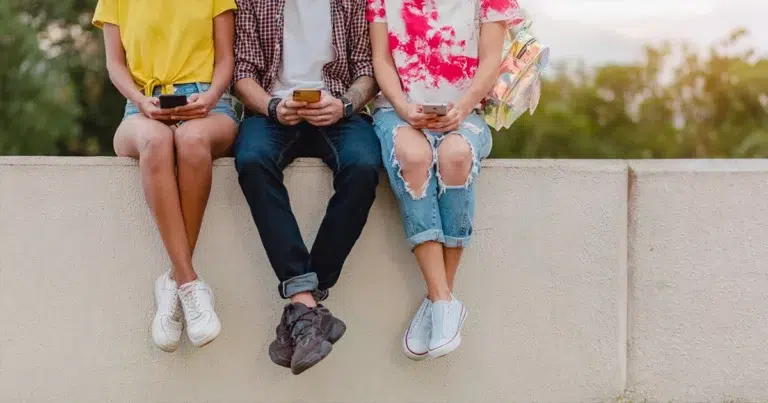 Generation Y nutzt Smartphone im Alltag. Drei junge Erwachsene sitzen nebeneinander auf einer Mauer und tauschen sich aus