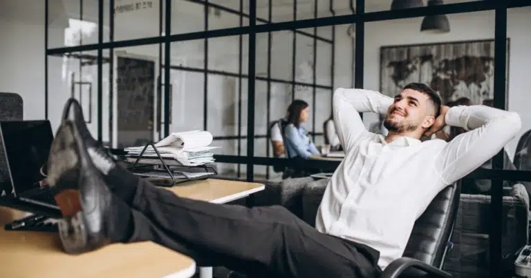 Mitarbeiter lehnt sich entspannt am Schreibtisch zurück während der Arbeitszeit und macht eine ausgiebige Pause.